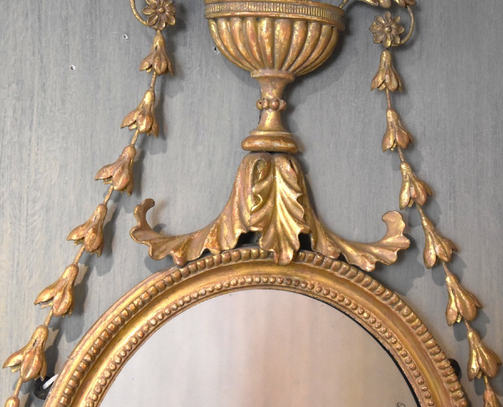 An oval Georgian giltwood mirror featuring a carved urn surmount filled with wheat sheaves, trailing floral husk swags, and an acanthus leaf bottom finial. View IV