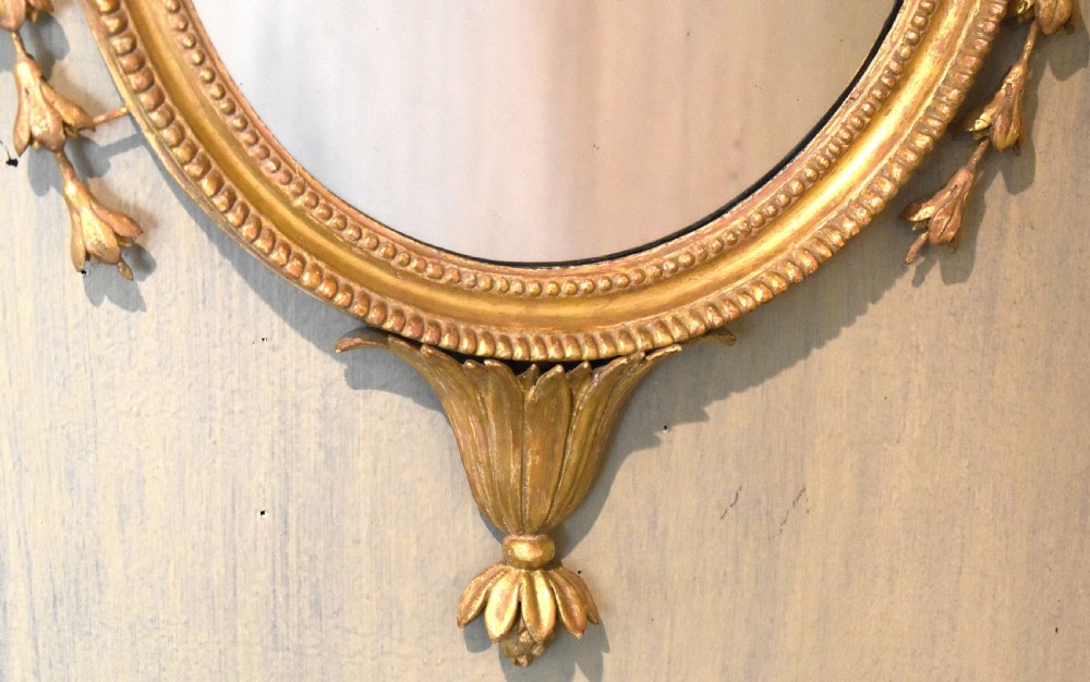 An oval Georgian giltwood mirror featuring a carved urn surmount filled with wheat sheaves, trailing floral husk swags, and an acanthus leaf bottom finial. View V