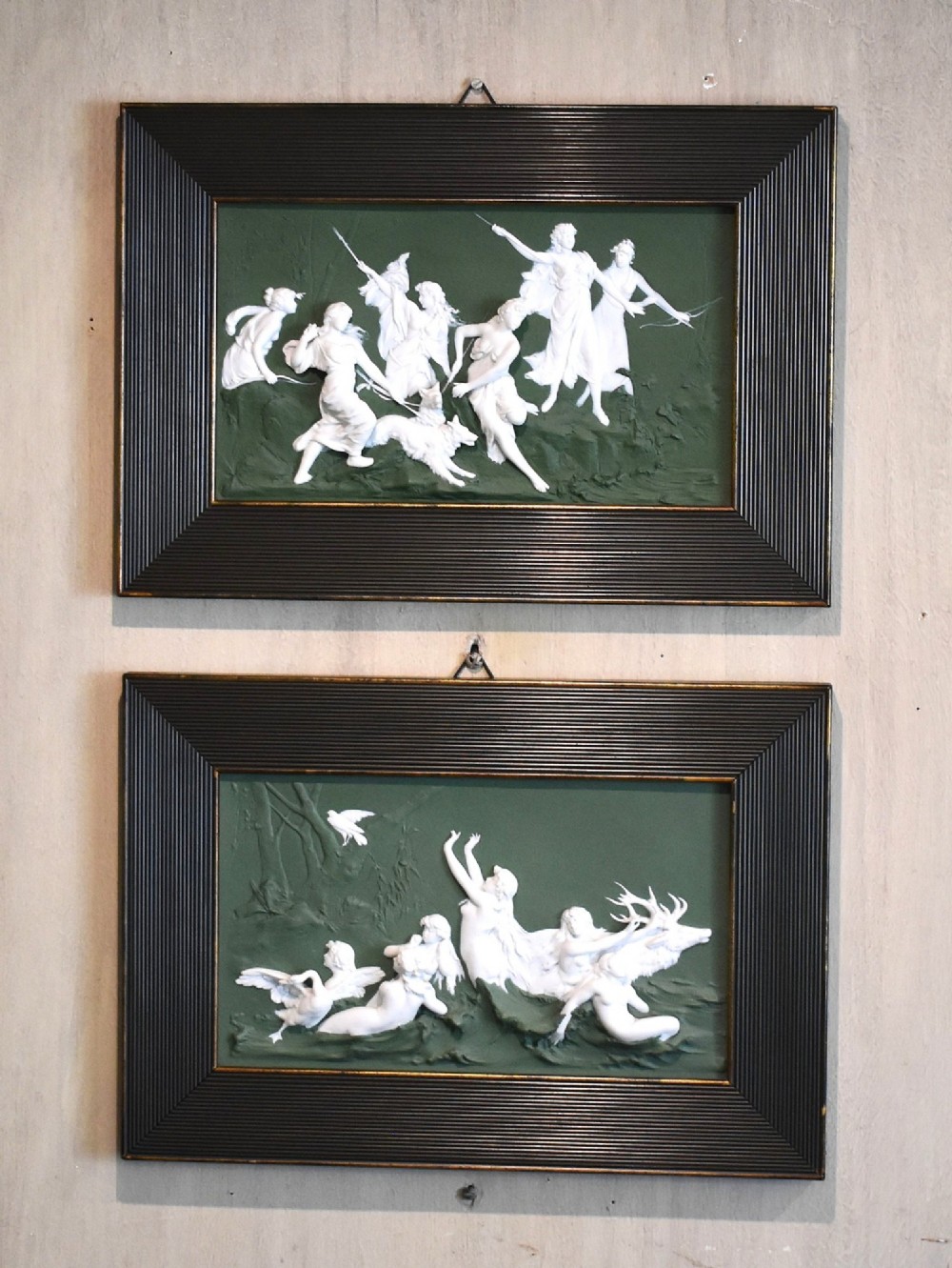 A pair of framed Art Nouveau Dresden porcelain plaques featuring white high-relief mythological figures on a dark green ground within reeded black frames.