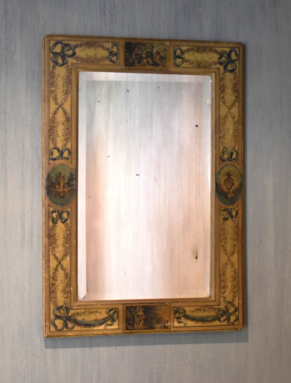 A rectangular wall mirror featuring a bevelled glass pane surrounded by a wide frame decorated with hand-painted silk motifs including ribbon bows, floral swags, and central medallions.