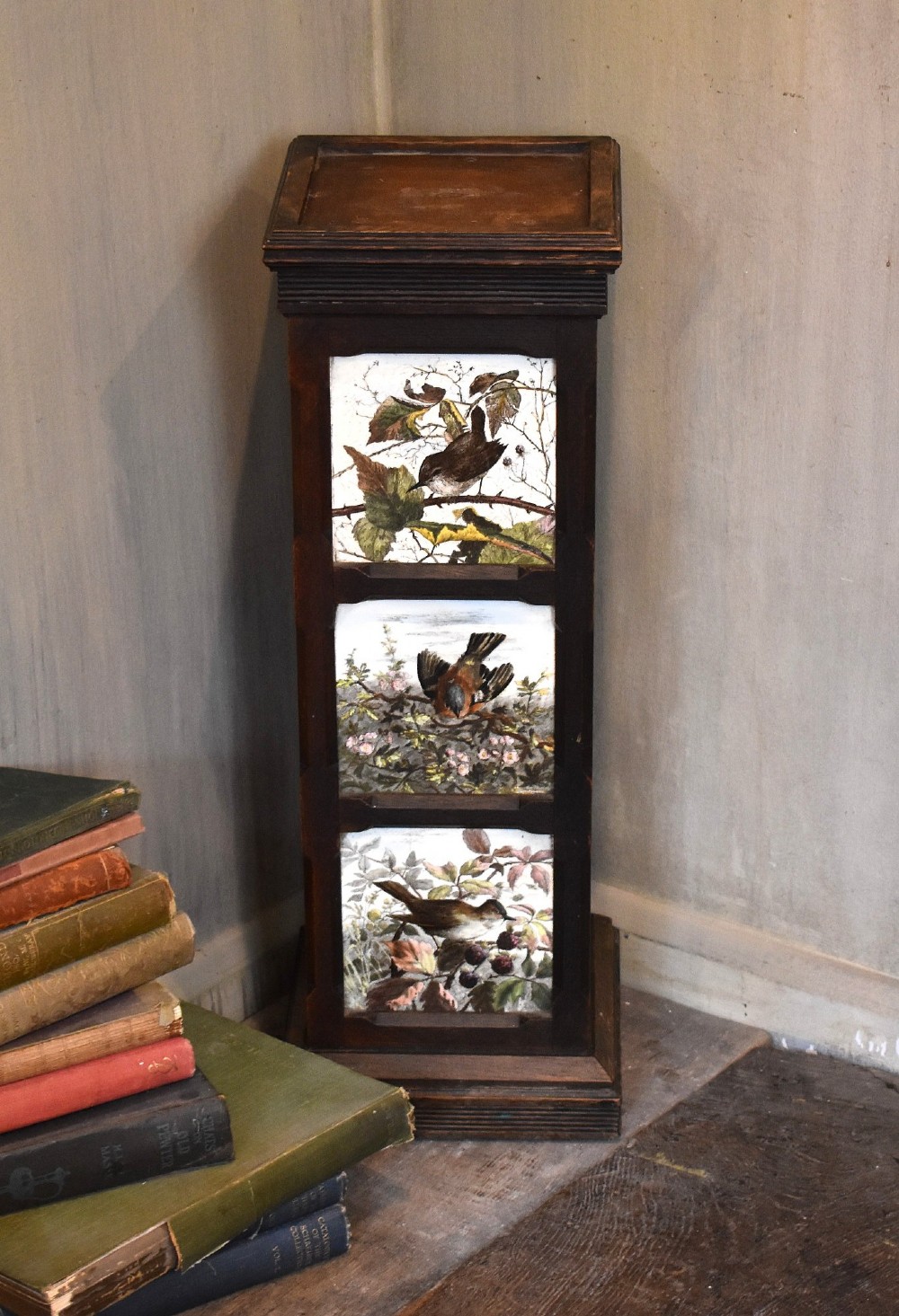 A dark wood Aesthetic Movement pedestal column with three vertical inset ceramic tiles displaying painted birds amidst foliage.