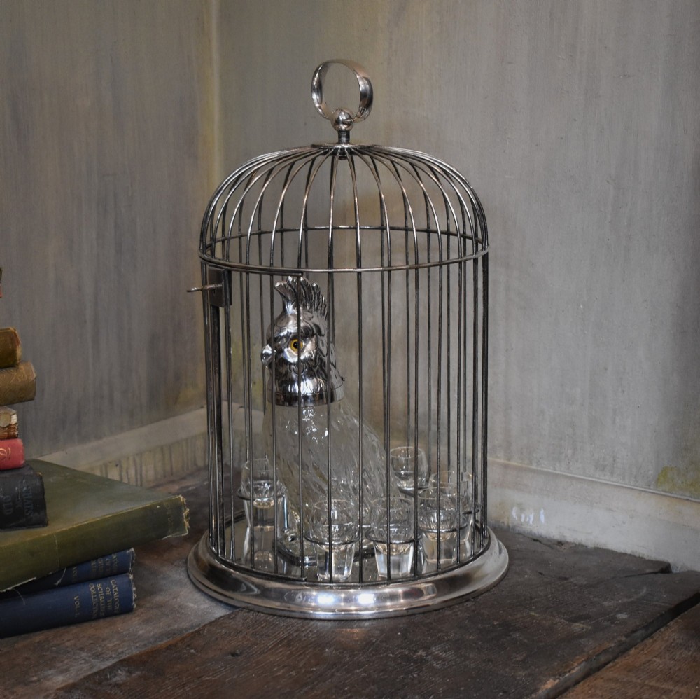 A silverplate birdcage tantalus containing a glass cockatoo-shaped decanter with a silverplate head stopper and surrounding shot glasses.