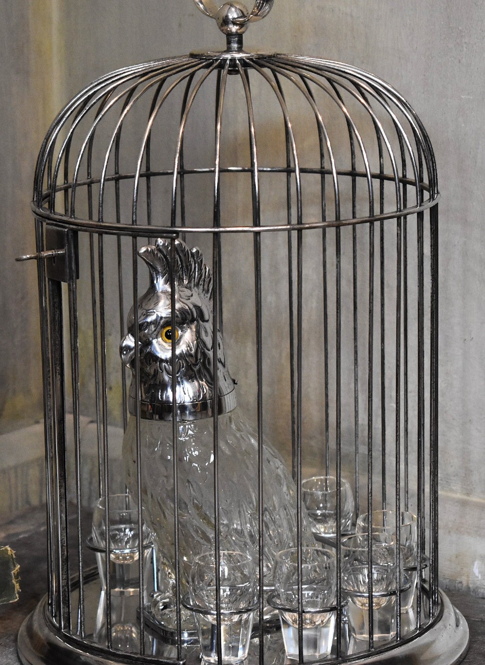 A silverplate birdcage tantalus containing a glass cockatoo-shaped decanter with a silverplate head stopper and surrounding shot glasses. View II