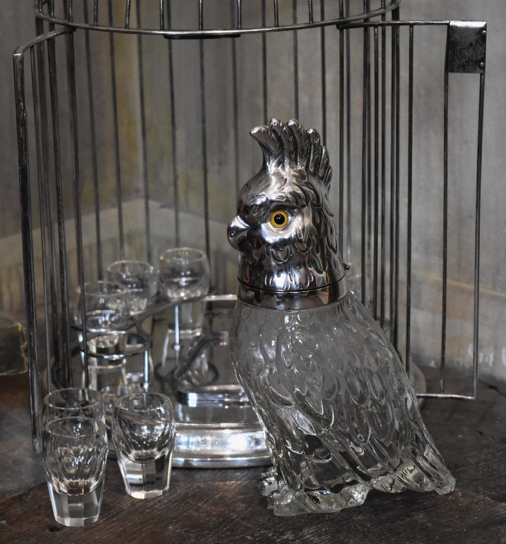 A silverplate birdcage tantalus containing a glass cockatoo-shaped decanter with a silverplate head stopper and surrounding shot glasses. View VI