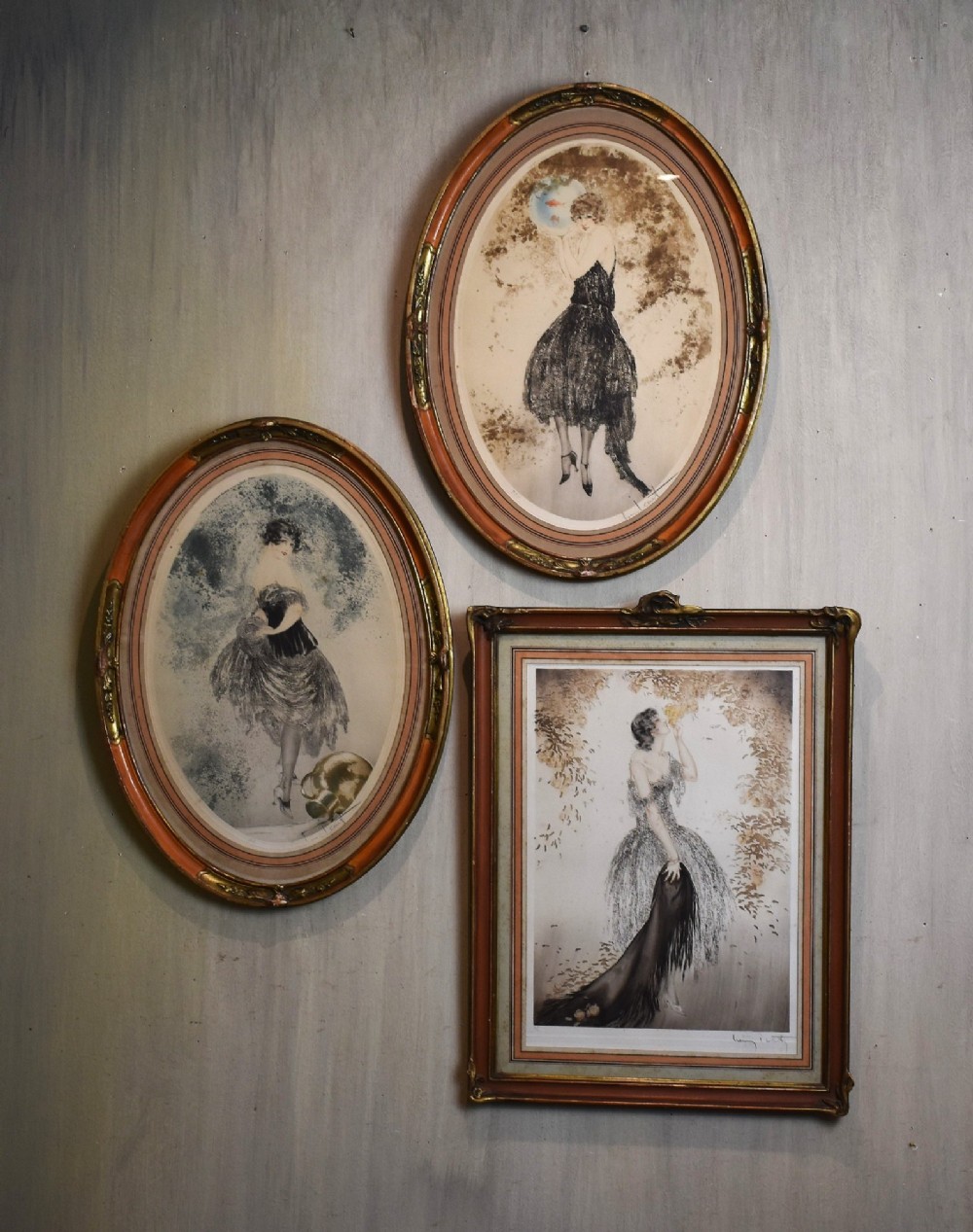 A set of three framed Art Deco fashion prints by Louis Icart featuring figures in 1920s gowns, presented in two oval and one rectangular gilt frames.