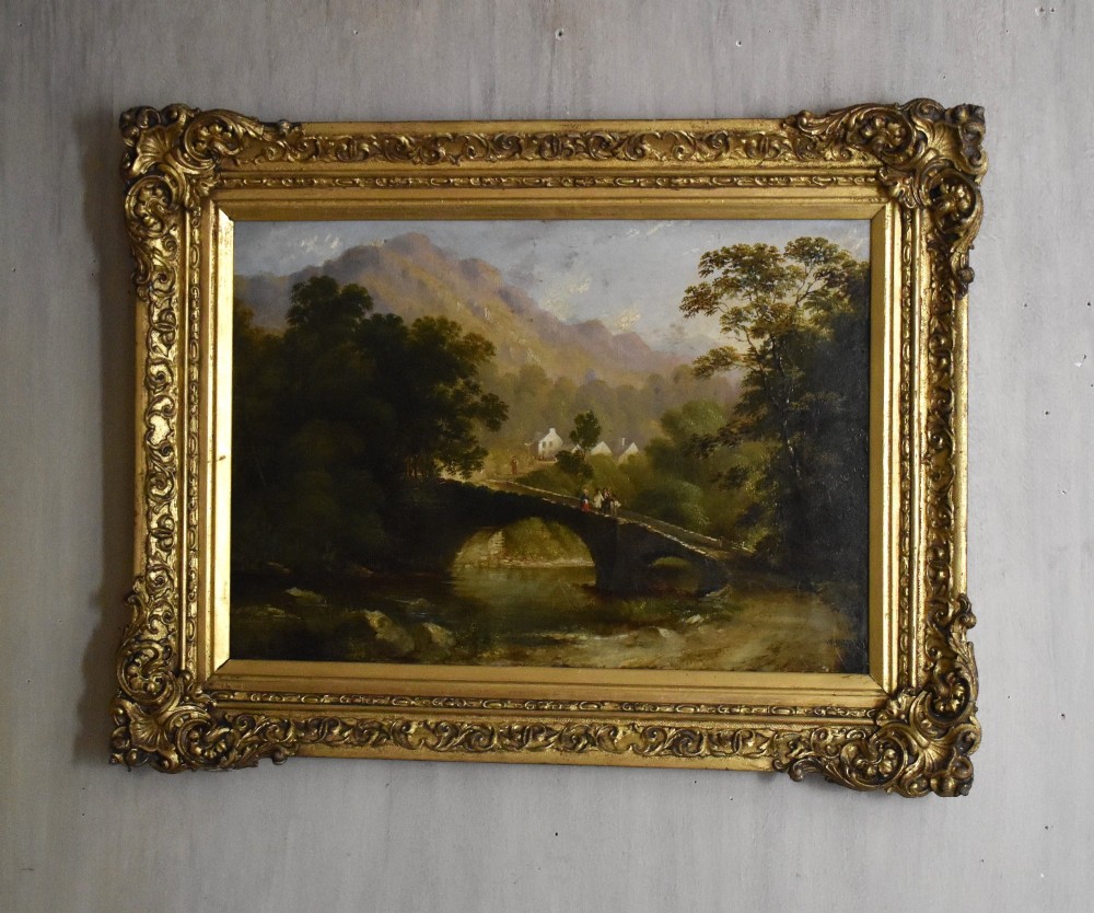 A nineteenth-century Victorian oil painting depicting a river scene with a stone arch bridge and distant mountains, housed within an ornate gilded frame.