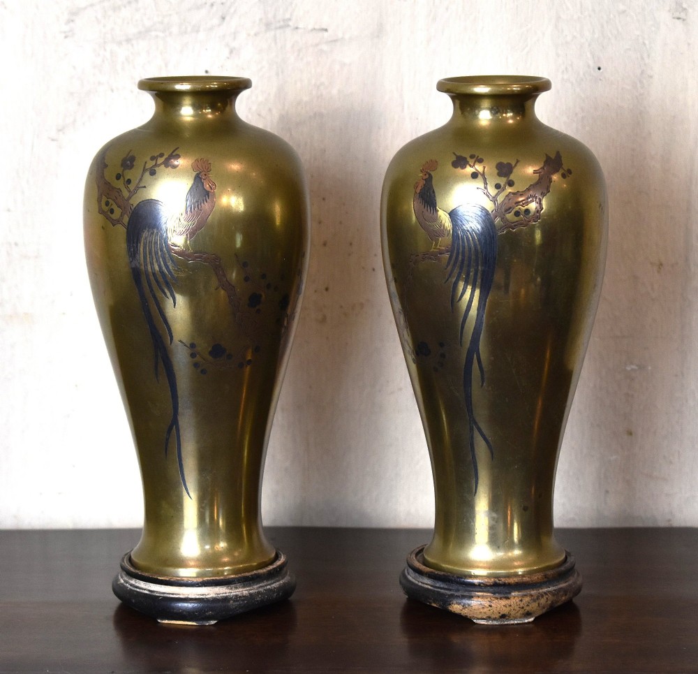 A pair of baluster-shaped golden bronze vases with detailed mixed-metal inlays depicting cockerels perched on branches, set on dark carved wooden bases.