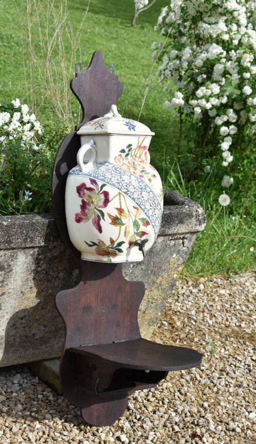 fig 1. An antique French Art Nouveau cream ceramic wall fountain decorated with botanical floral transfers and a spigot, mounted on a dark carved wooden backboard with a small shelf.