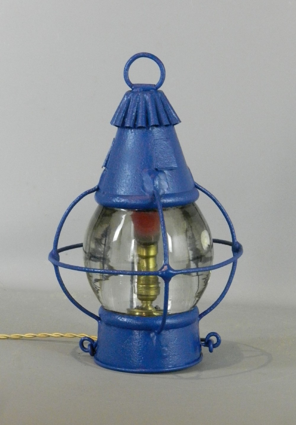 A blue painted metal ships globe lantern with a central clear glass shade, protective wire cage, top ring handle, and electrical wiring.