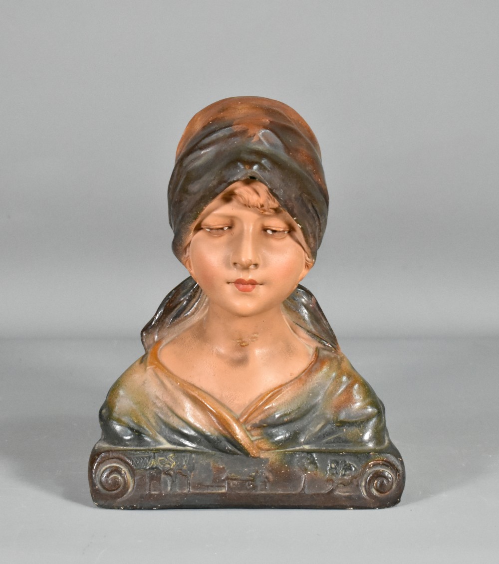 A painted plaster portrait bust depicting a young girl wearing a draped headscarf, resting on a scrolled base.