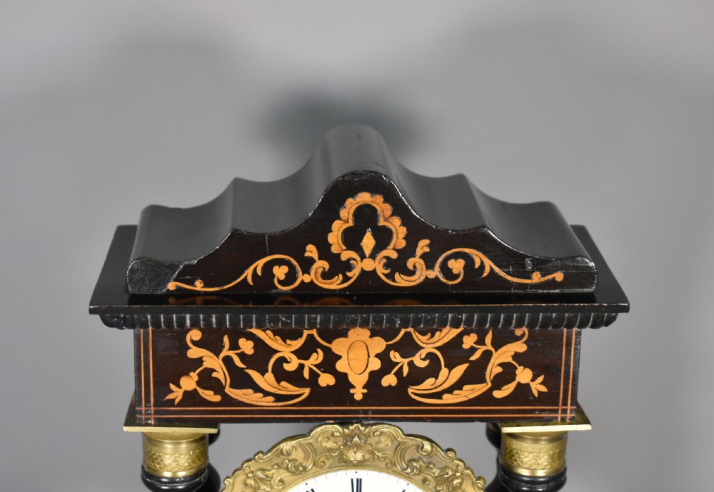An ebonised Napoleon III portico mantel clock featuring floral marquetry inlays, twin barley twist columns, a white enamel dial with Roman numerals, and gilt brass mounts. View V