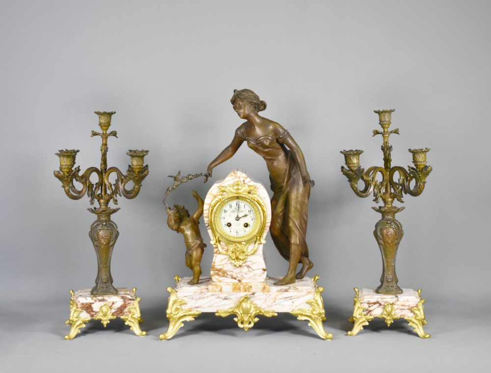 An Art Nouveau French mantel clock garniture featuring a central sculptural bronze figure beside an enamel clock face mounted on a pink veined marble base, flanked by two matching multi-arm candelabra.