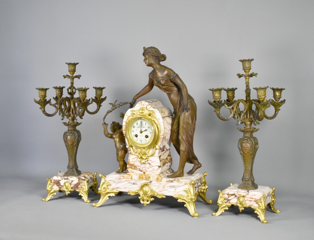 An Art Nouveau French mantel clock garniture featuring a central sculptural bronze figure beside an enamel clock face mounted on a pink veined marble base, flanked by two matching multi-arm candelabra. View III