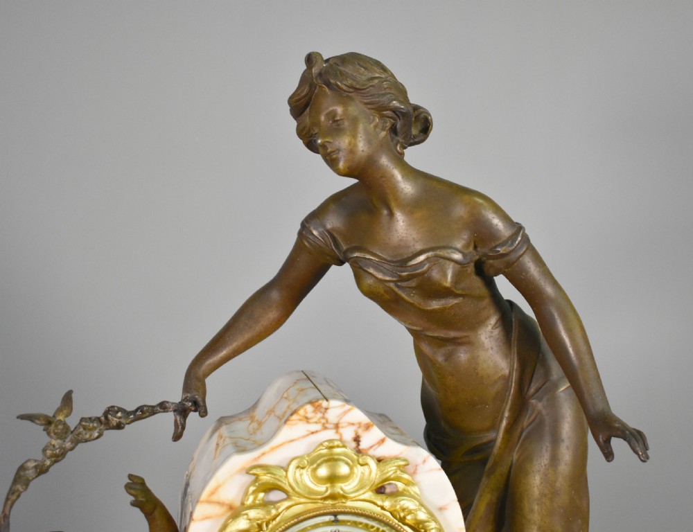 An Art Nouveau French mantel clock garniture featuring a central sculptural bronze figure beside an enamel clock face mounted on a pink veined marble base, flanked by two matching multi-arm candelabra. View X