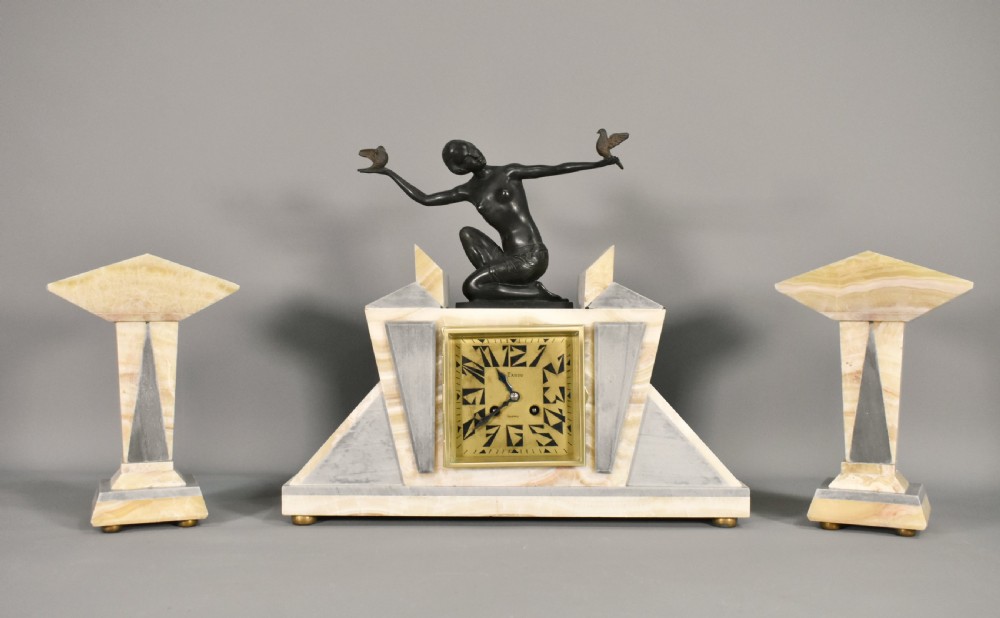 A three-piece French Art Deco clock garniture set featuring a central geometric onyx and marble clock topped with a bronze-finished figure of a female with birds, accompanied by two matching side pedestals.
