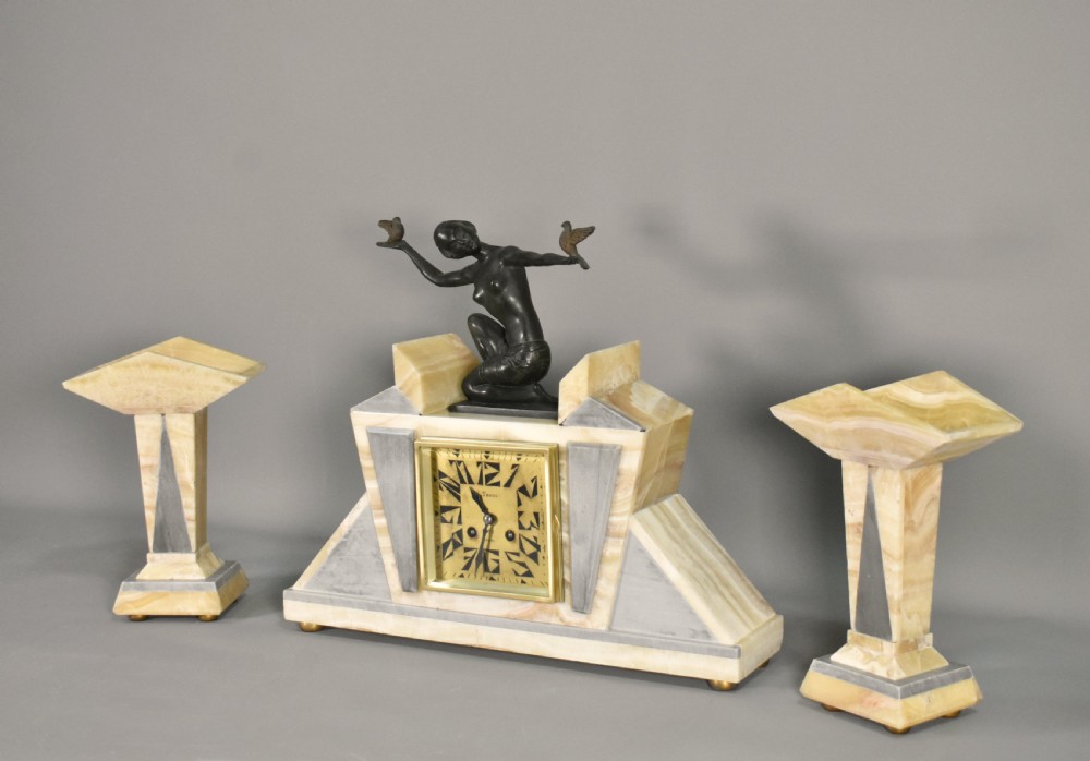 A three-piece French Art Deco clock garniture set featuring a central geometric onyx and marble clock topped with a bronze-finished figure of a female with birds, accompanied by two matching side pedestals. View V