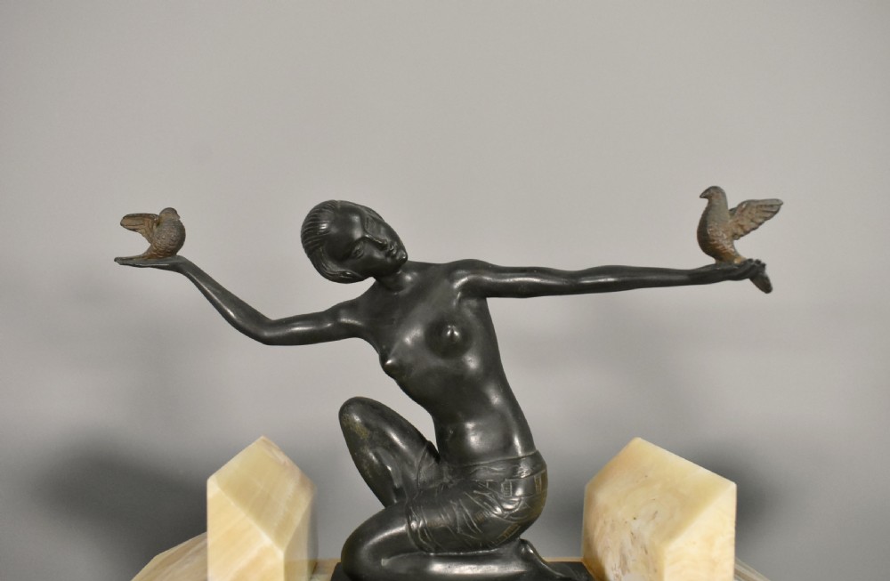 A three-piece French Art Deco clock garniture set featuring a central geometric onyx and marble clock topped with a bronze-finished figure of a female with birds, accompanied by two matching side pedestals. View VI