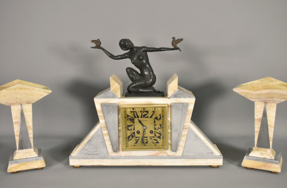 A three-piece French Art Deco clock garniture set featuring a central geometric onyx and marble clock topped with a bronze-finished figure of a female with birds, accompanied by two matching side pedestals. View XVIII
