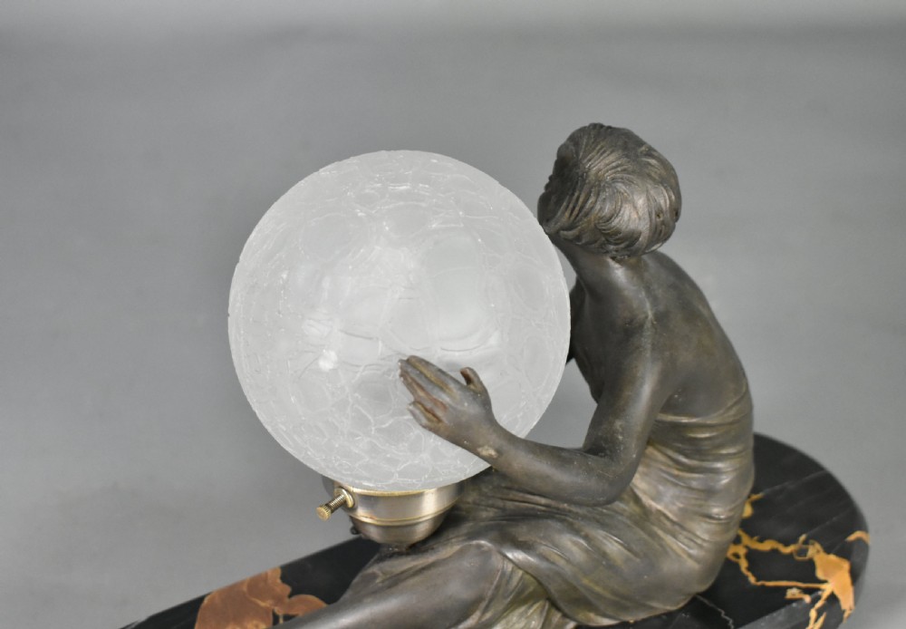 An Art Deco table lamp featuring a dark patinated female figure seated beside a spherical textured glass shade on an oval black marble base with yellow veining. View VII