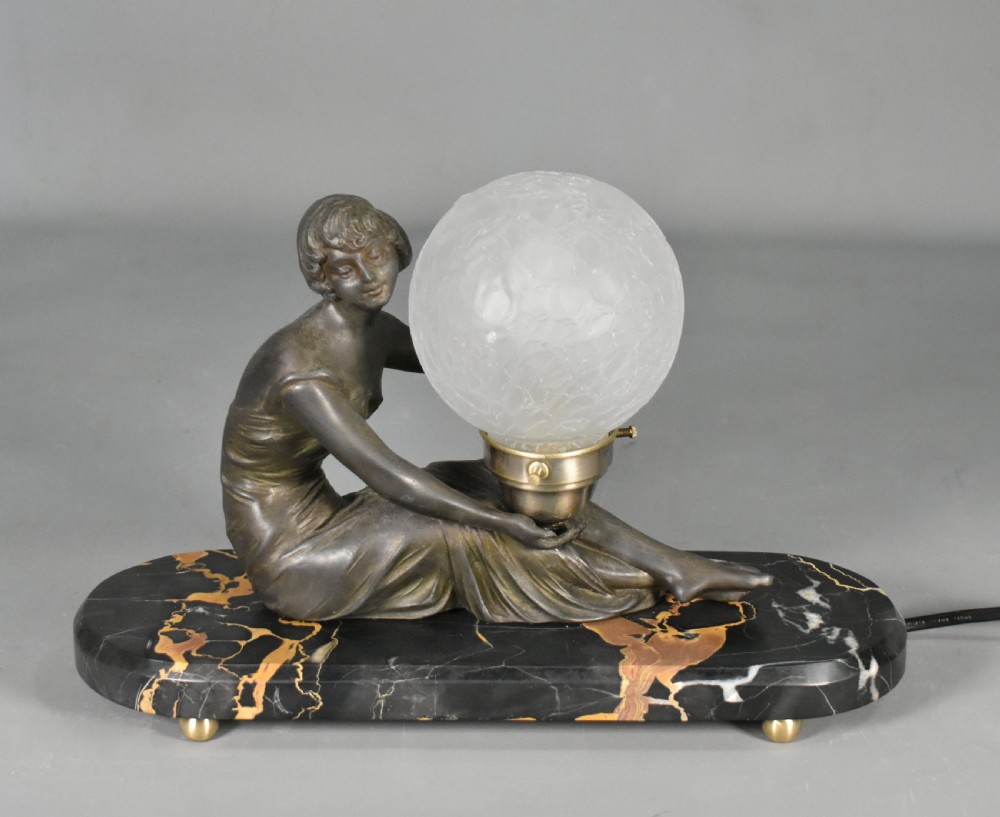 An Art Deco table lamp featuring a dark patinated female figure seated beside a spherical textured glass shade on an oval black marble base with yellow veining. View X