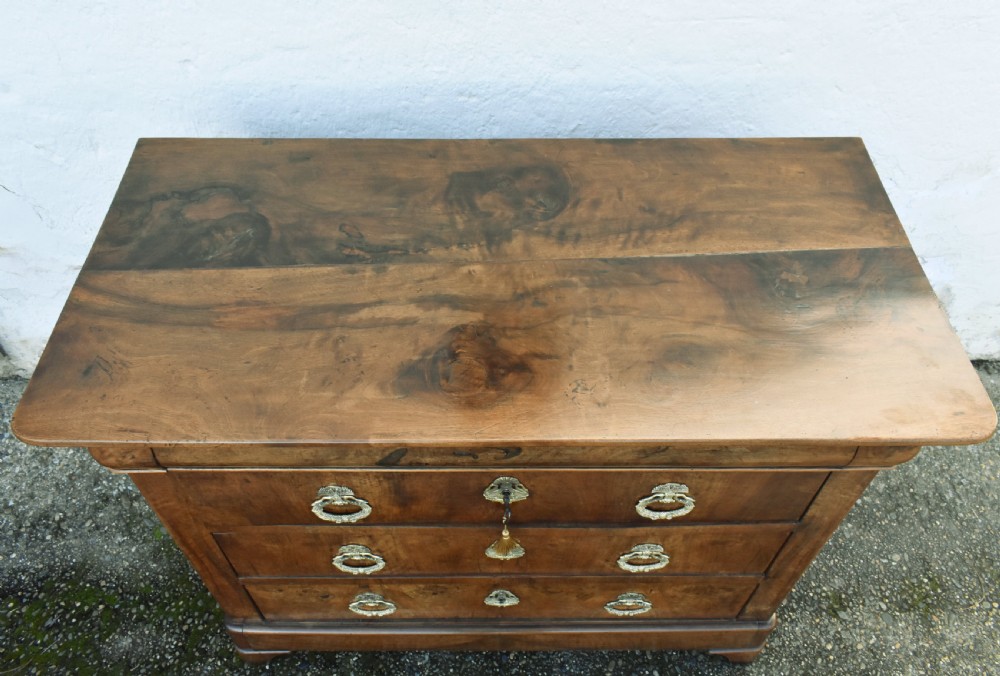 A 19th-century French Louis Philippe walnut commode featuring a rounded top drawer, three lower drawers with brass drop handles, and a plinth base. View X