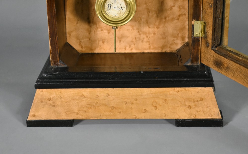 An antique mantel clock featuring a birdseye maple veneered arched case, ebonised plinth base, white enamel dial with Roman numerals, and a brass pendulum. View X