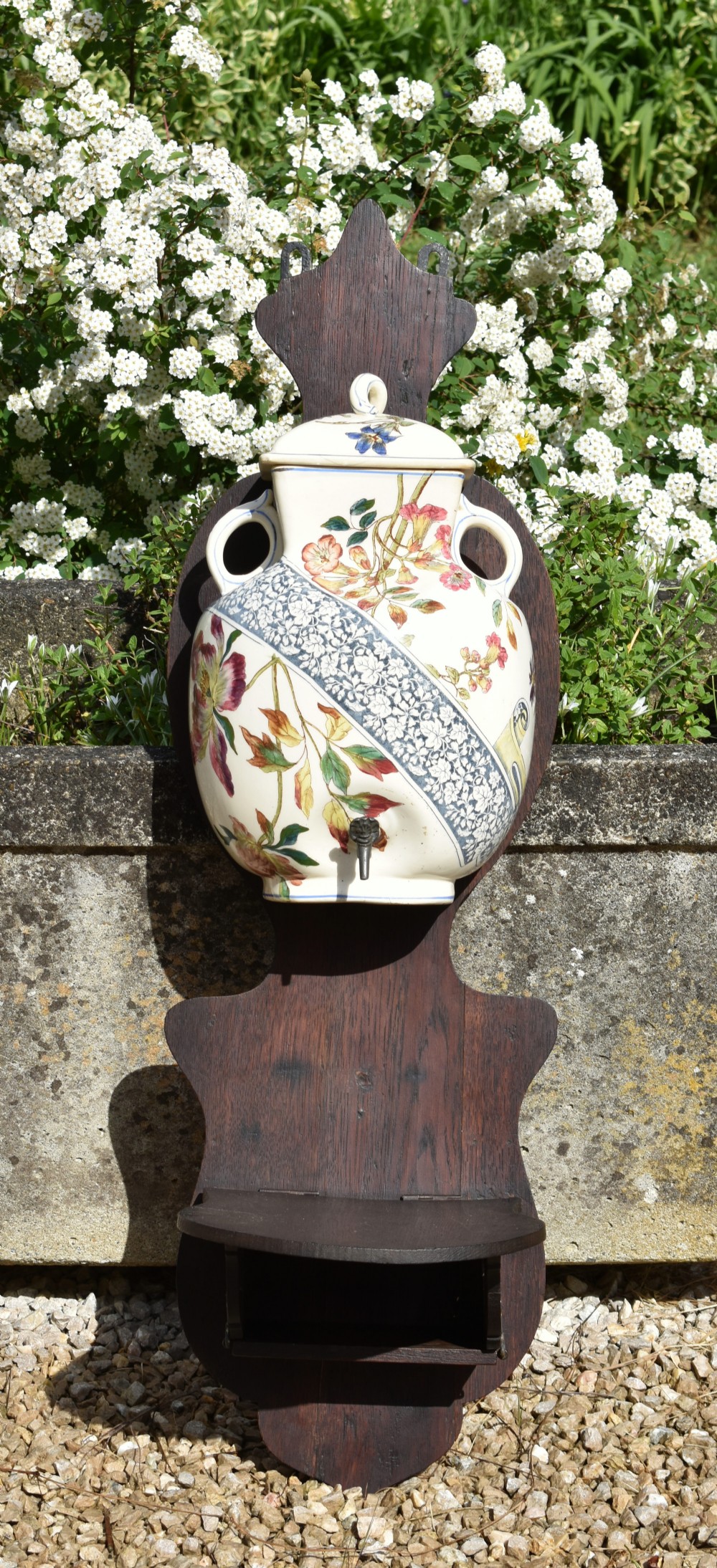 An antique French Art Nouveau cream ceramic wall fountain decorated with botanical floral transfers and a spigot, mounted on a dark carved wooden backboard with a small shelf.