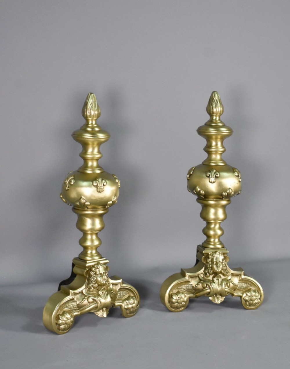 A pair of cast bronze Louis XIV style firedogs featuring baluster shafts, flame finials, and scrolled bases with relief mask carvings. View II