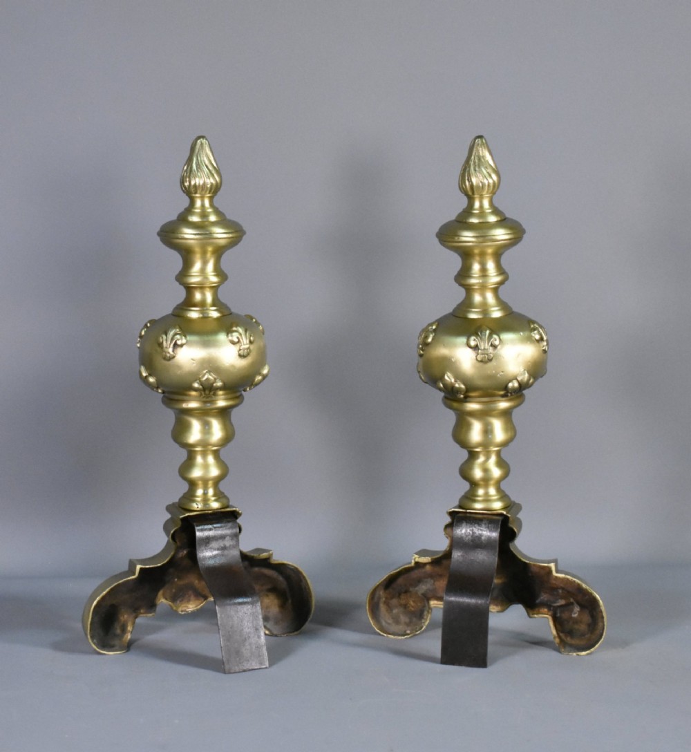 A pair of cast bronze Louis XIV style firedogs featuring baluster shafts, flame finials, and scrolled bases with relief mask carvings. View X