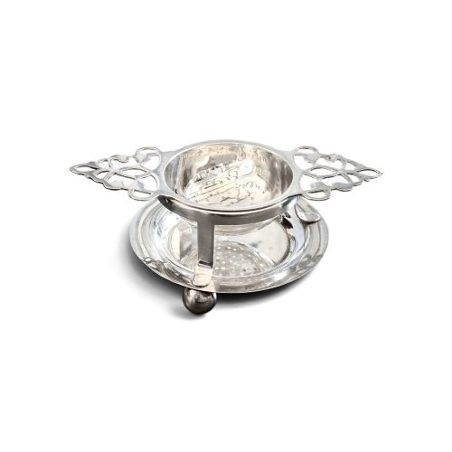 fig 1. A sterling silver tea strainer featuring openwork pierced handles resting on a matching circular drip stand supported by spherical ball feet.