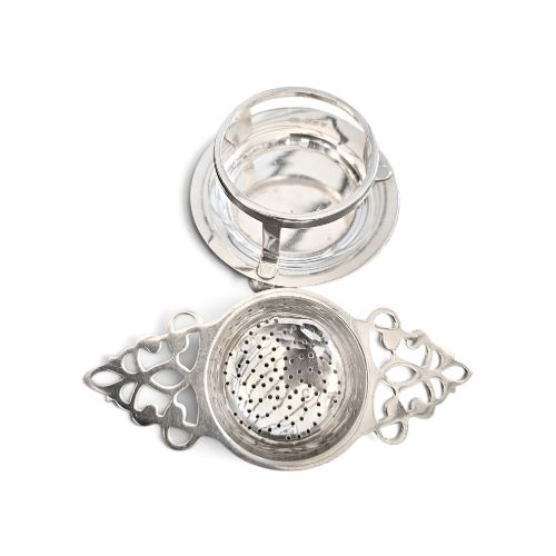 fig 2. A sterling silver tea strainer featuring openwork pierced handles resting on a matching circular drip stand supported by spherical ball feet.