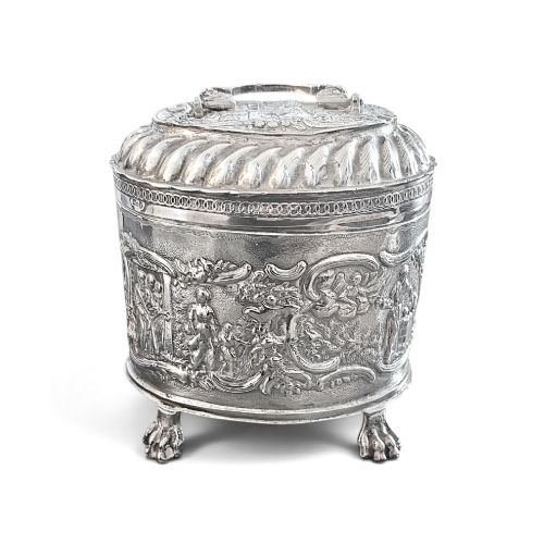 fig 1. An oval sterling silver tea caddy featuring repousse figurative scenes, a hinged lid with handle, and four claw feet.