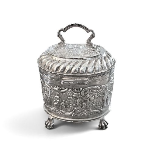 fig 2. An oval sterling silver tea caddy featuring repousse figurative scenes, a hinged lid with handle, and four claw feet.