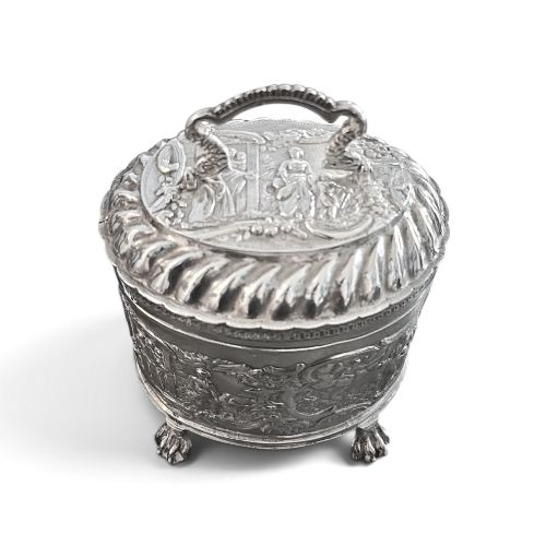 fig 3. An oval sterling silver tea caddy featuring repousse figurative scenes, a hinged lid with handle, and four claw feet.