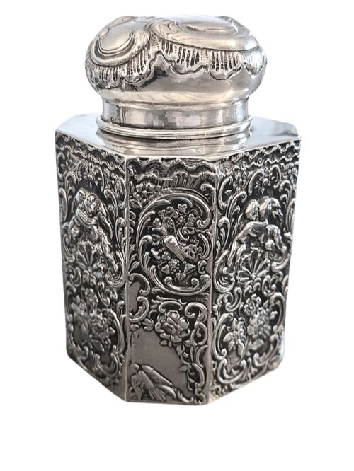 fig 1. A late Victorian hexagonal sterling silver tea caddy featuring relief repousse work of cherubs, scrollwork, and floral motifs with a fitted domed lid.