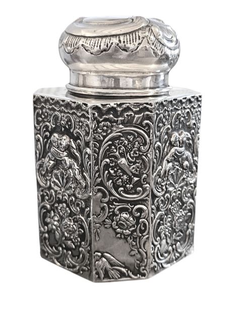 fig 2. A late Victorian hexagonal sterling silver tea caddy featuring relief repousse work of cherubs, scrollwork, and floral motifs with a fitted domed lid.