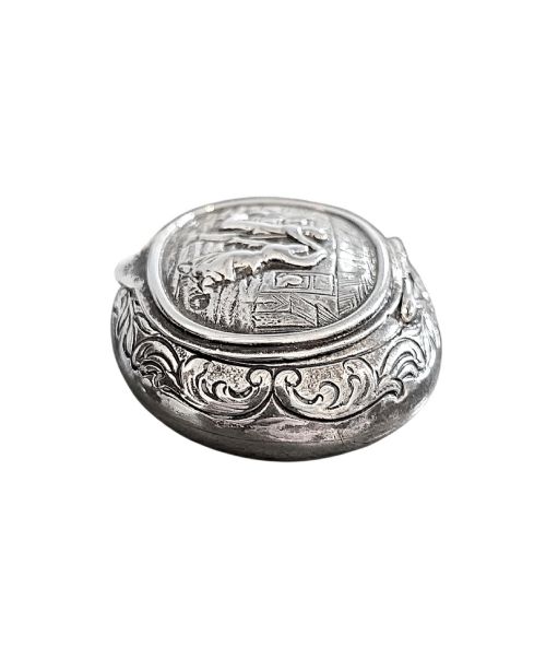fig 2. An oval antique sterling silver snuff box featuring an embossed central scene of dancing figures, framed by scrollwork borders.