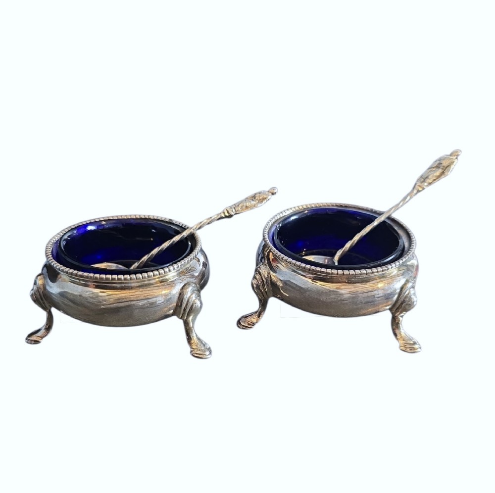 A pair of Victorian sterling silver open salt cellars featuring beaded rims, cabriole legs, cobalt blue glass liners, and matching silver salt spoons.