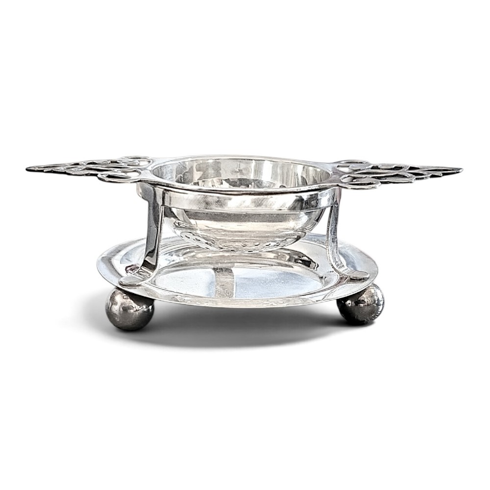 A sterling silver tea strainer featuring openwork pierced handles resting on a matching circular drip stand supported by spherical ball feet.