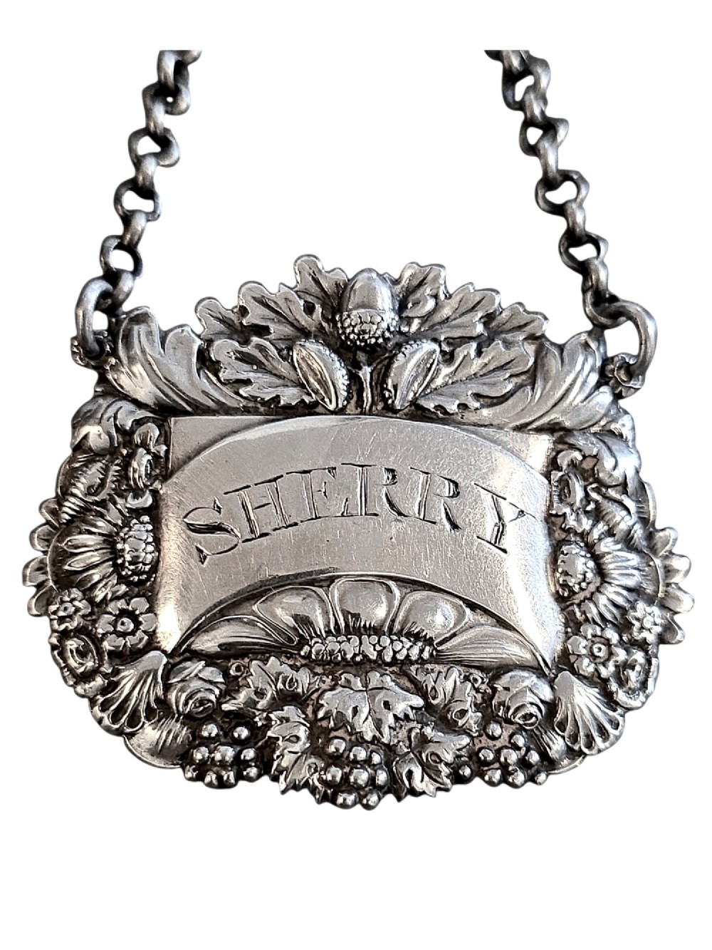 A Georgian sterling silver sherry decanter label featuring ornate floral and acorn repousse borders, an engraved central banner, and a chain suspension.
