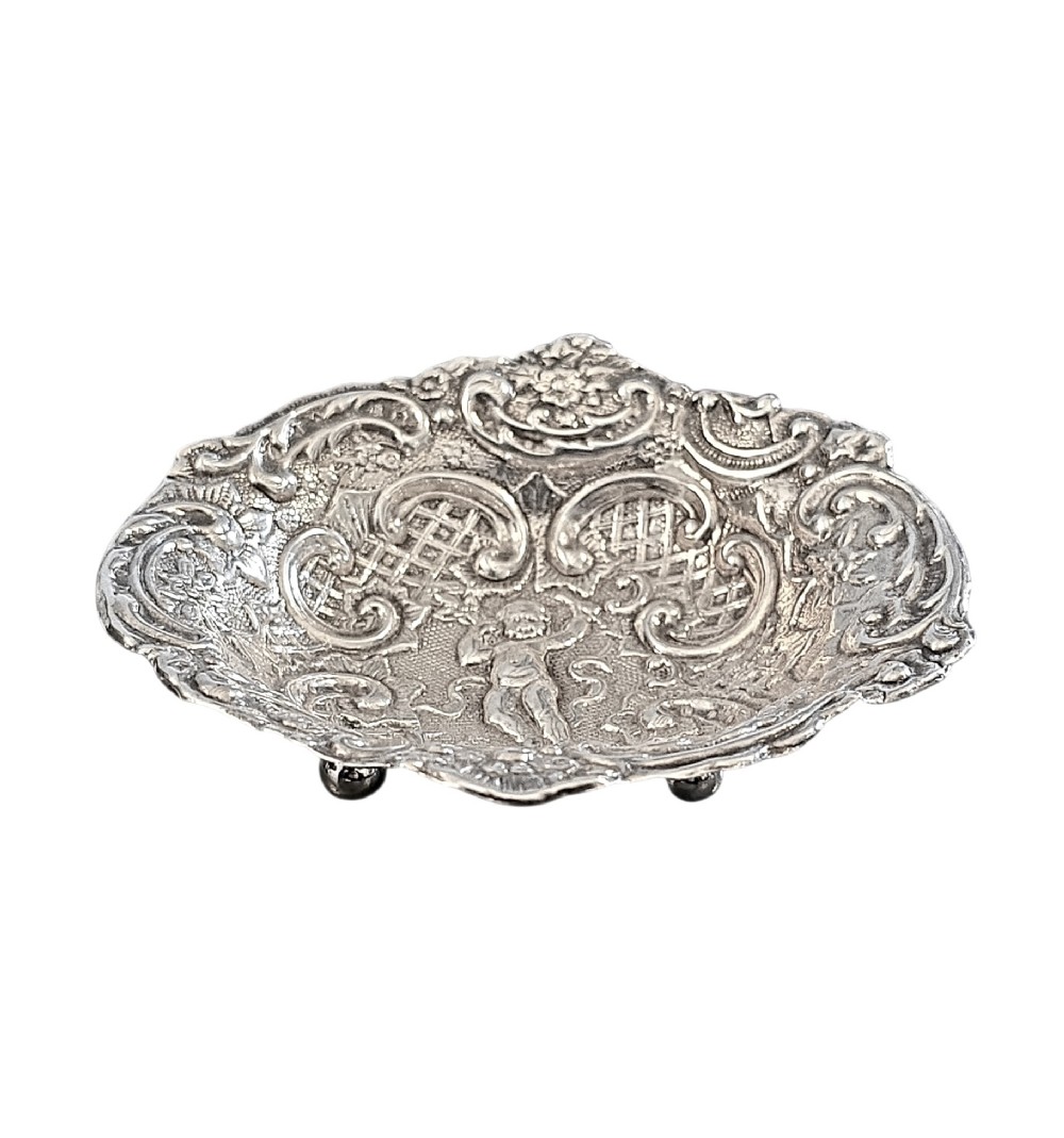 An antique oval sterling silver bonbon dish featuring embossed scrollwork, lattice detailing, a central cherub figure, and spherical ball feet.