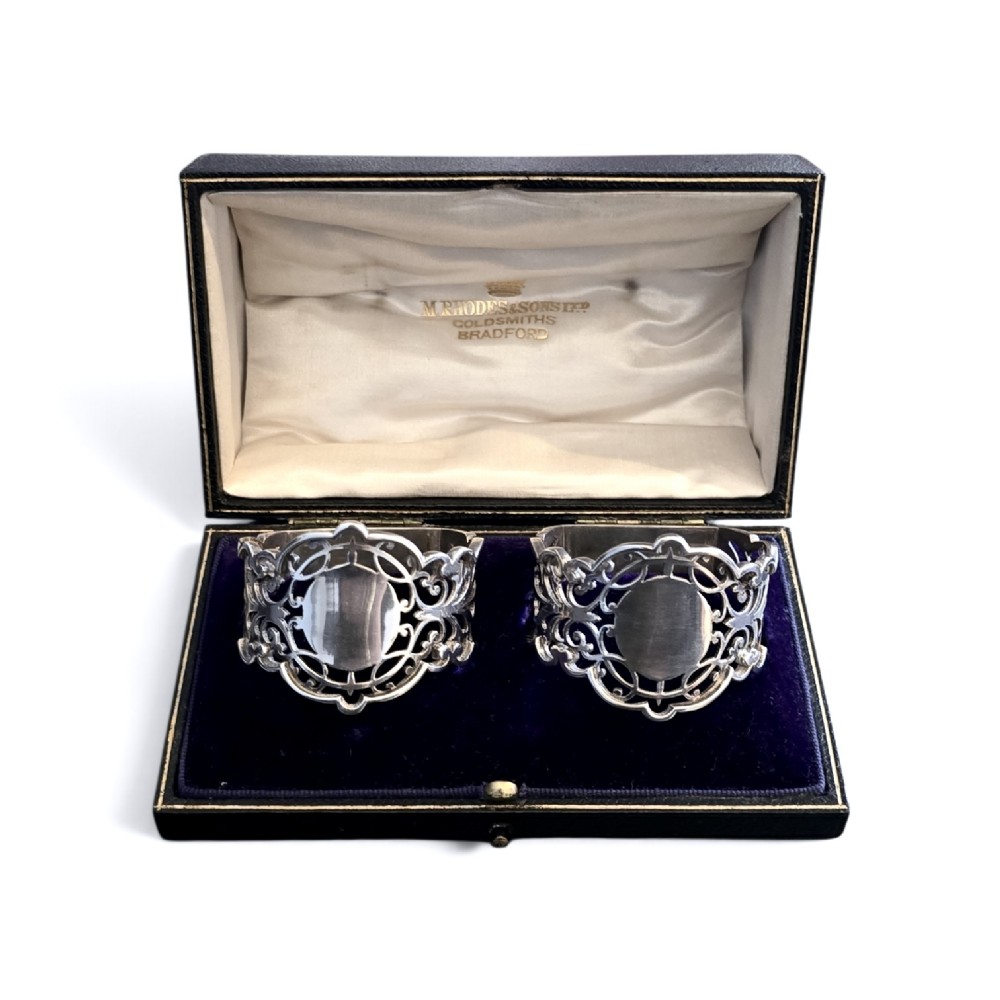 A pair of Edwardian pierced sterling silver napkin rings with unengraved cartouches presented in a dark velvet-lined fitted box.