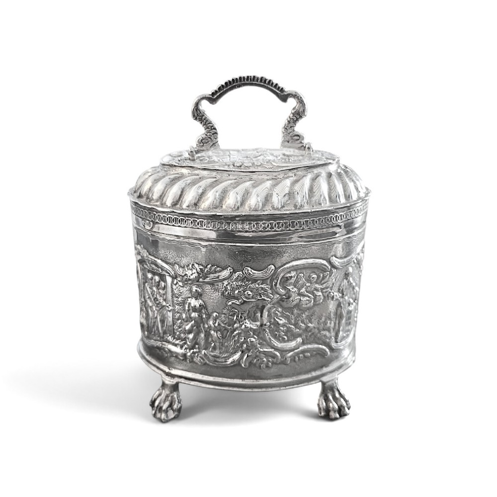 An oval sterling silver tea caddy featuring repousse figurative scenes, a hinged lid with handle, and four claw feet.