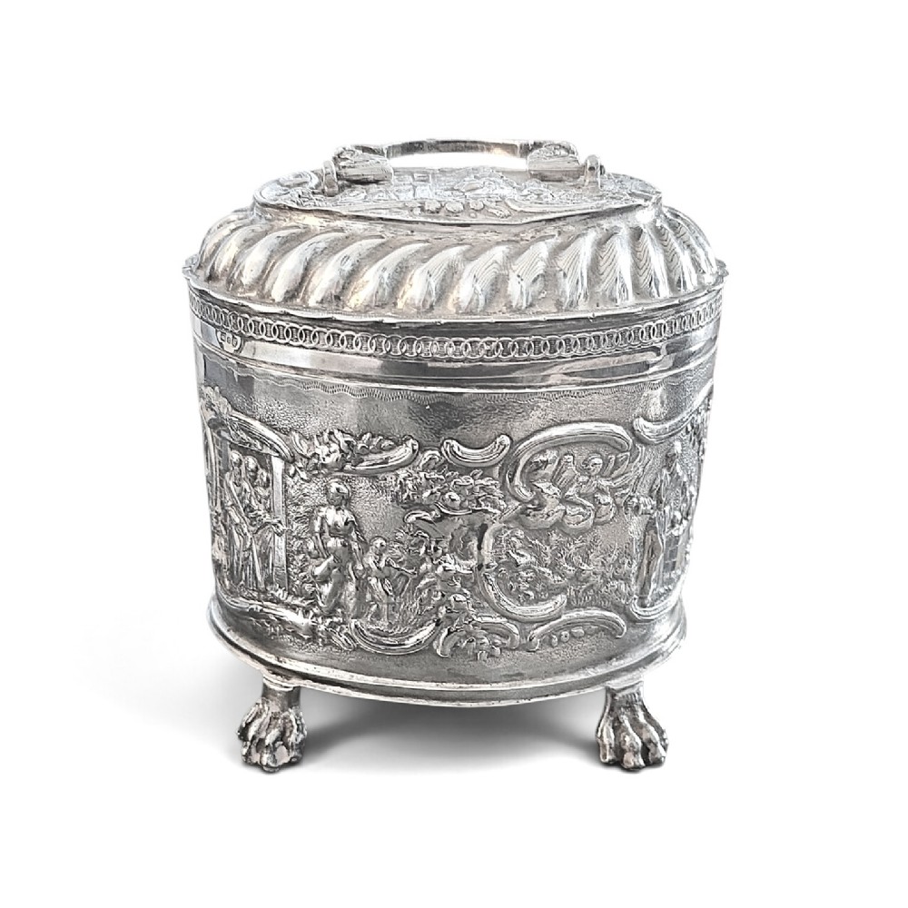 An oval sterling silver tea caddy featuring repousse figurative scenes, a hinged lid with handle, and four claw feet. View II