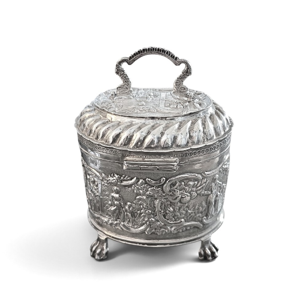 An oval sterling silver tea caddy featuring repousse figurative scenes, a hinged lid with handle, and four claw feet. View III