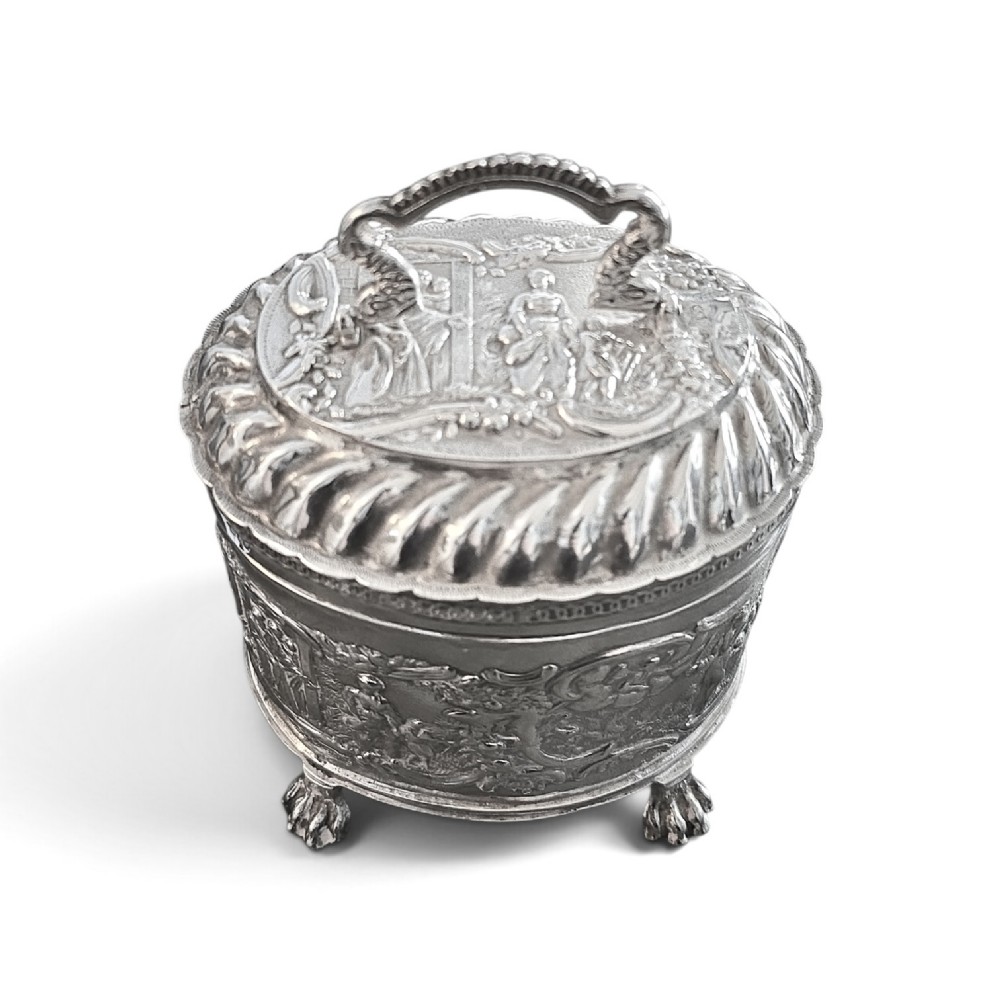 An oval sterling silver tea caddy featuring repousse figurative scenes, a hinged lid with handle, and four claw feet. View IV