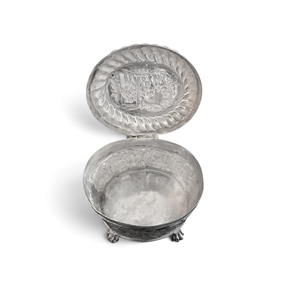 An oval sterling silver tea caddy featuring repousse figurative scenes, a hinged lid with handle, and four claw feet. View V