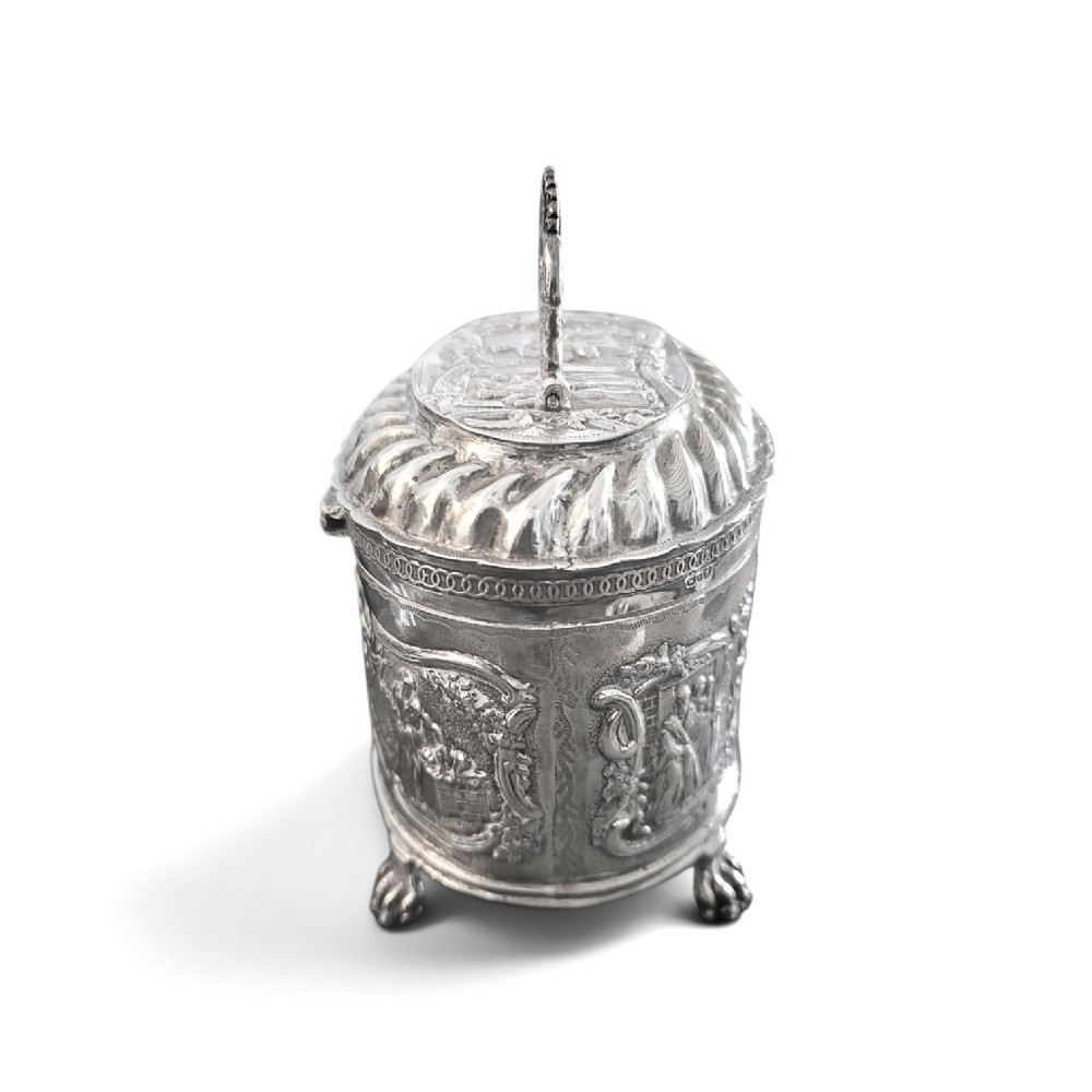 An oval sterling silver tea caddy featuring repousse figurative scenes, a hinged lid with handle, and four claw feet. View VI