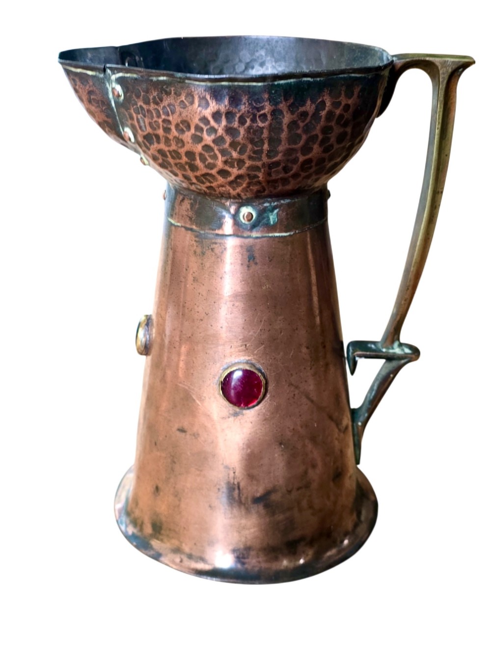 An antique Arts and Crafts copper jug featuring a planished finish, riveted collar, brass handle, and inset coloured cabochons.