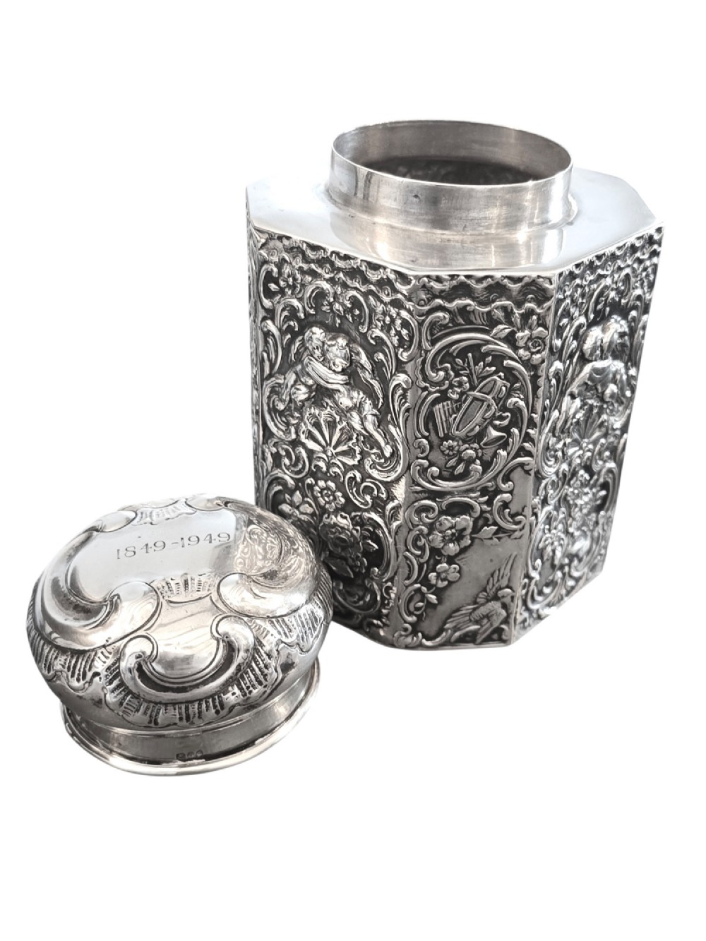 A late Victorian hexagonal sterling silver tea caddy featuring relief repousse work of cherubs, scrollwork, and floral motifs with a fitted domed lid. View IV